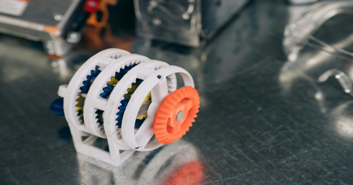 A table displays various 3D printed objects including a vase, a gear, and a miniature figure.