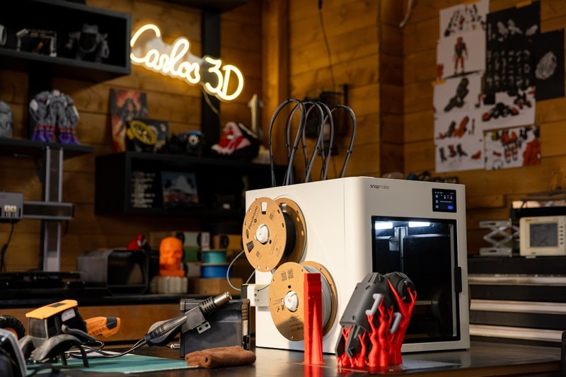 Collection of 3D printer accessories including filament dryer, build plates, and post-processing tools on a workbench